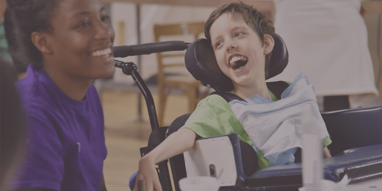 Young boy in a wheelchair smiles as he holds hands with his support staff