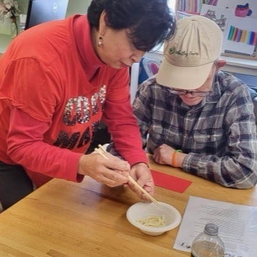 A staff member helps a person supported with Day Services practice using chopsticks for Lunar New Year.
